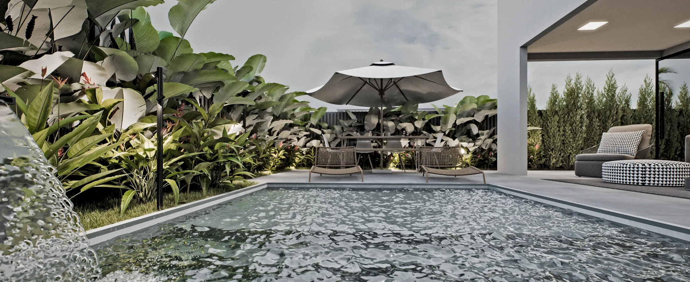 Piscina retangular com lâmina d'água na parede, borda de plantas tropicais, guarda-sol com mesa e um estar coberto com sofá à direita.